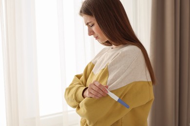 Photo of Upset woman with negative pregnancy test near window at home, space for text