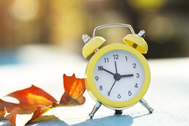 Photo of Autumn time. Alarm clock and golden leaf on light surface outdoors, closeup