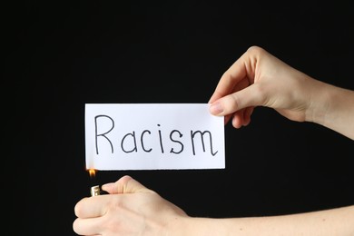 Photo of Woman lighting card with word Racism on black background, closeup