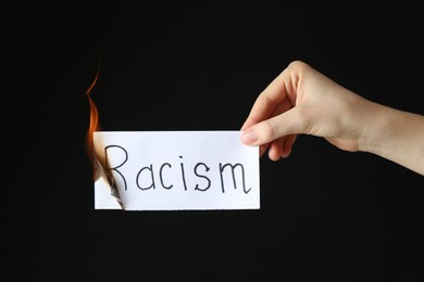 Photo of Woman holding burning card with word Racism on black background, closeup