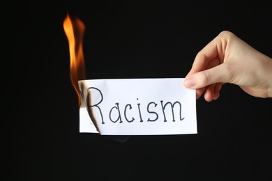 Photo of Woman holding burning card with word Racism on black background, closeup