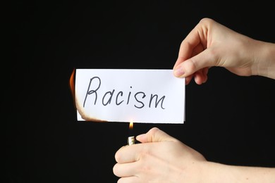 Photo of Woman lighting card with word Racism on black background, closeup