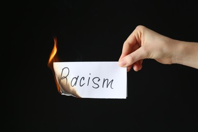 Photo of Woman holding burning card with word Racism on black background, closeup