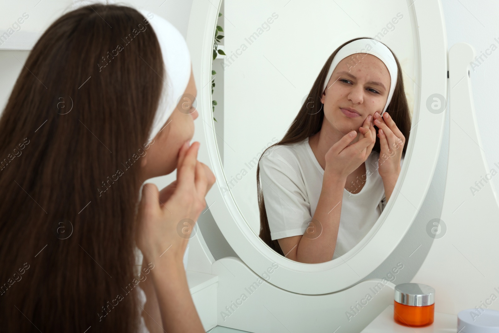 Photo of Upset teenage girl popping pimple on her face near mirror indoors. Acne problem