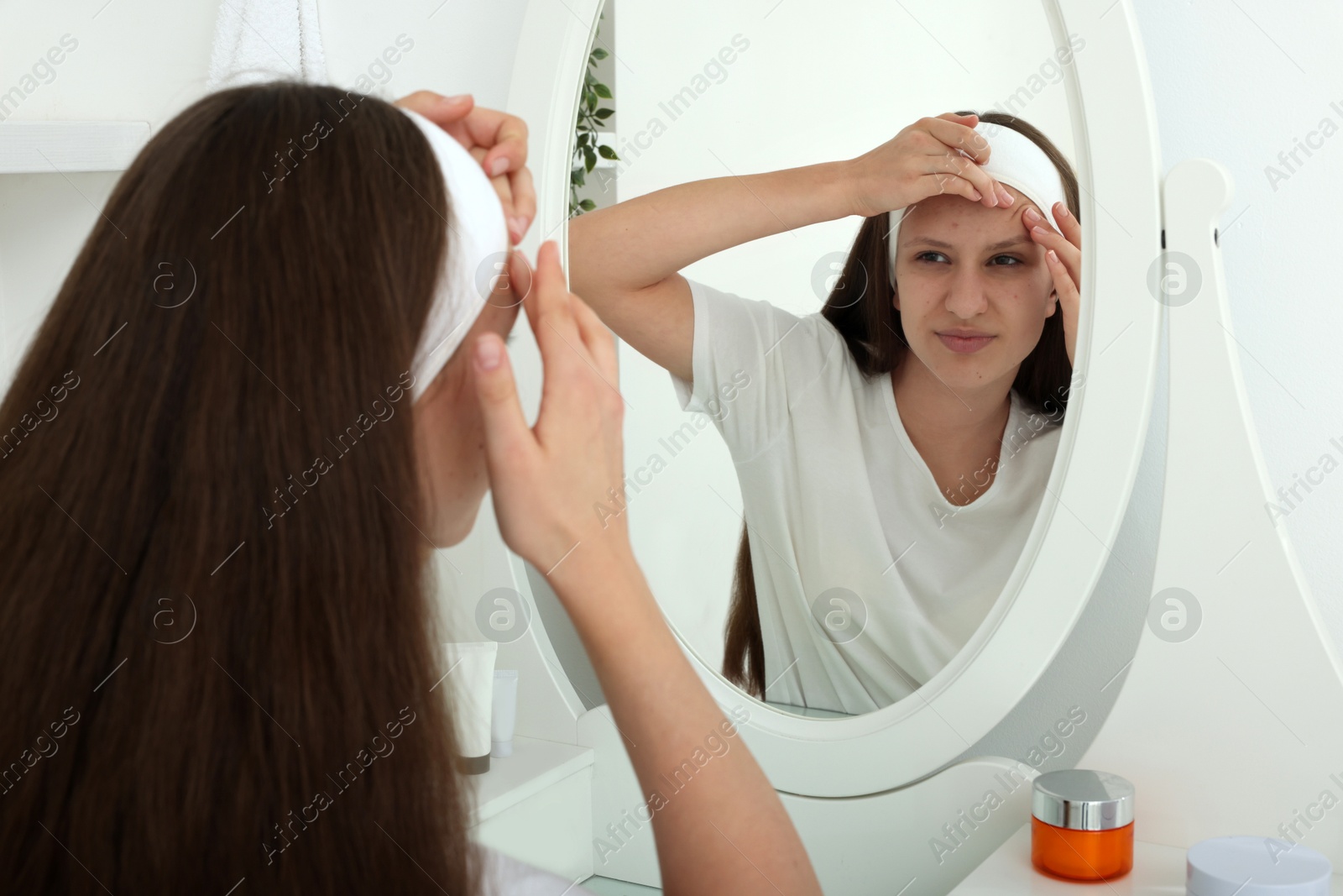 Photo of Upset teenage girl popping pimple on her face near mirror indoors. Acne problem