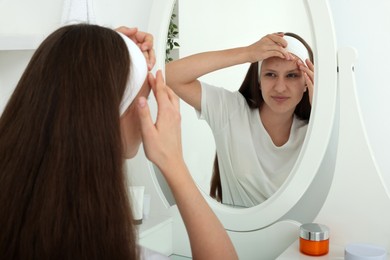 Photo of Upset teenage girl popping pimple on her face near mirror indoors. Acne problem