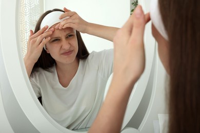 Photo of Upset teenage girl popping pimple on her face near mirror indoors. Acne problem