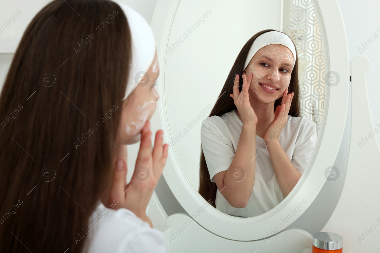 Photo of Teenage girl applying cream onto face near mirror indoors. Acne treatment