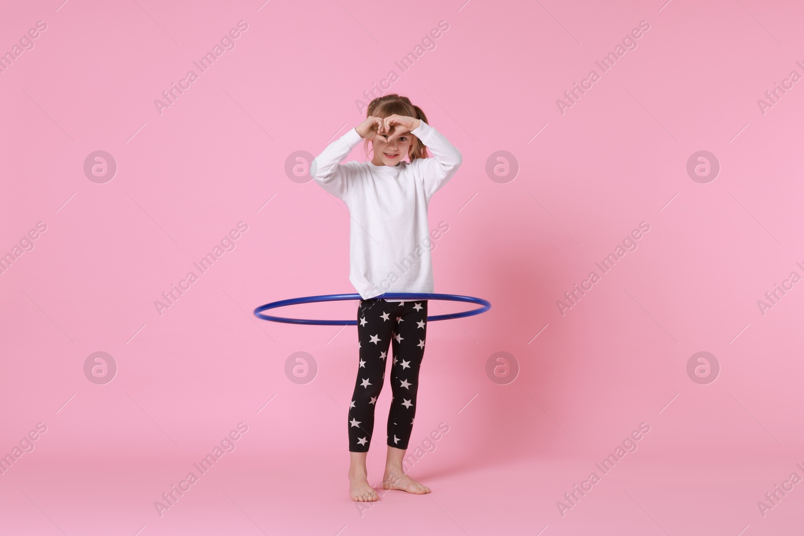 Photo of Cute little girl exercising with hula hoop on pink background