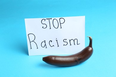 Photo of Card with phrase Stop Racism and dark banana with sad face on light blue background