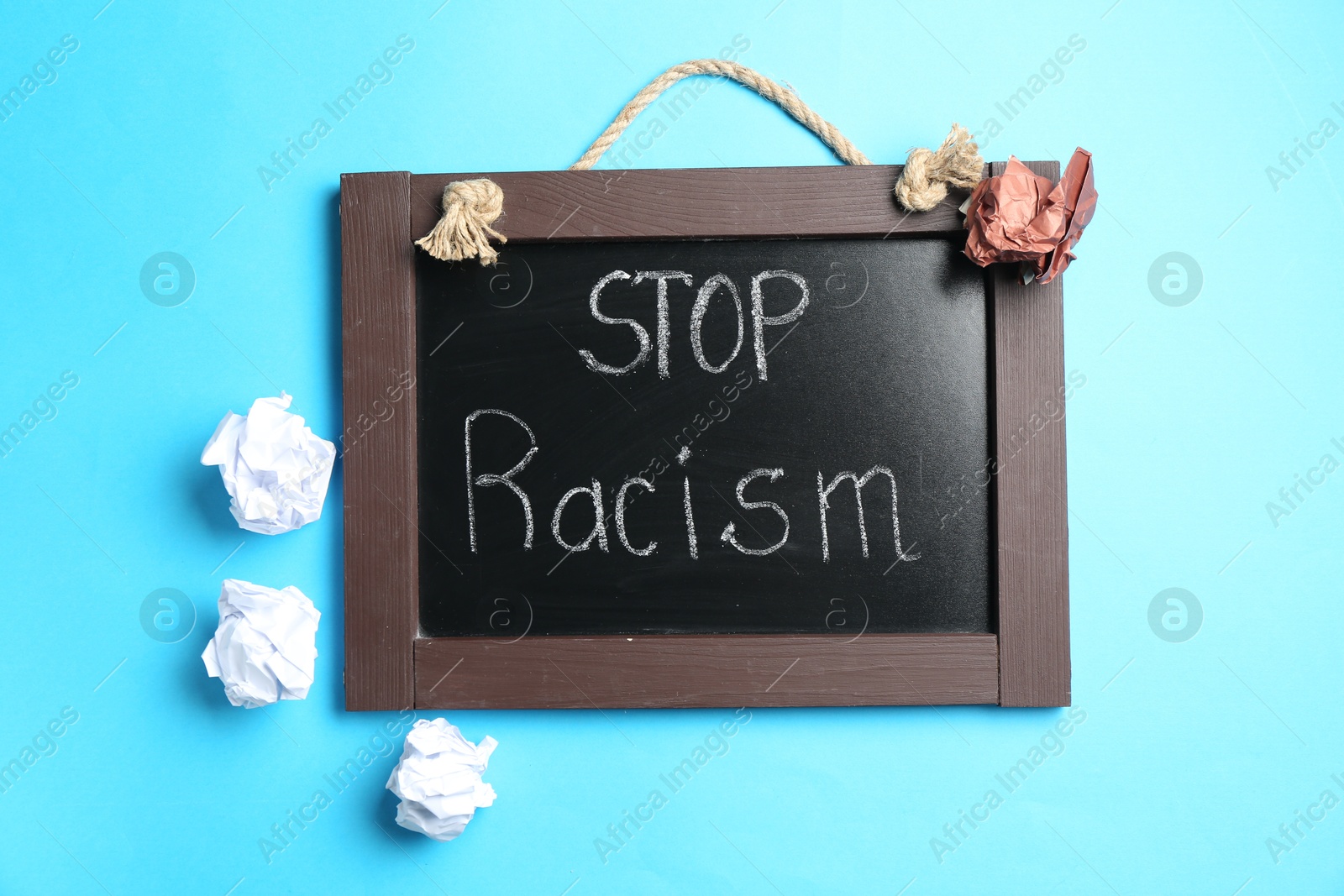 Photo of Chalkboard with phrase Stop Racism and crumpled paper pieces of different colors on light blue background, top view