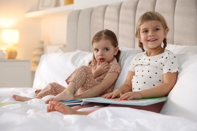 Cute little sisters reading book together in bed at home