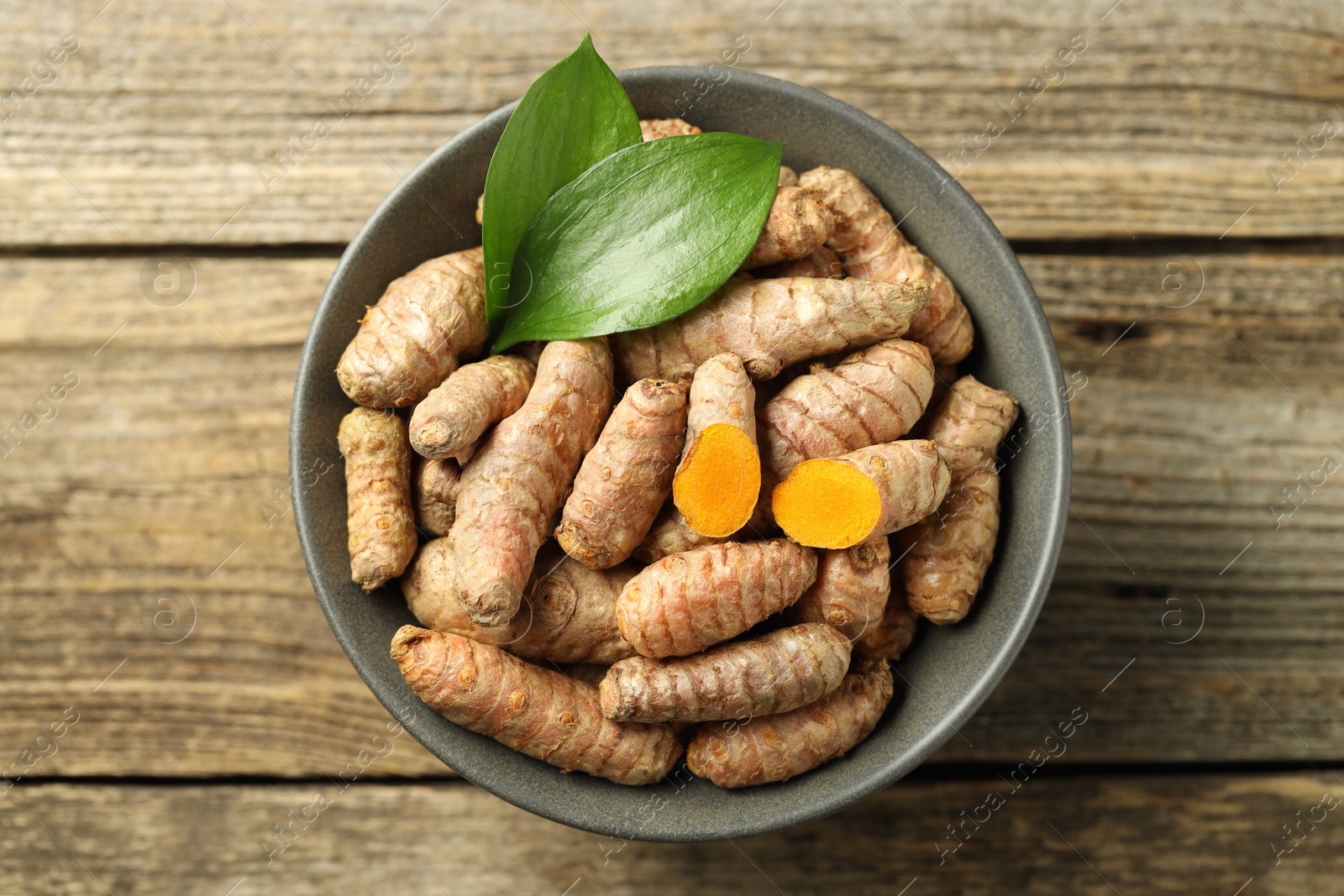 Photo of Tumeric rhizomes with leaves in bowl on wooden table, top view