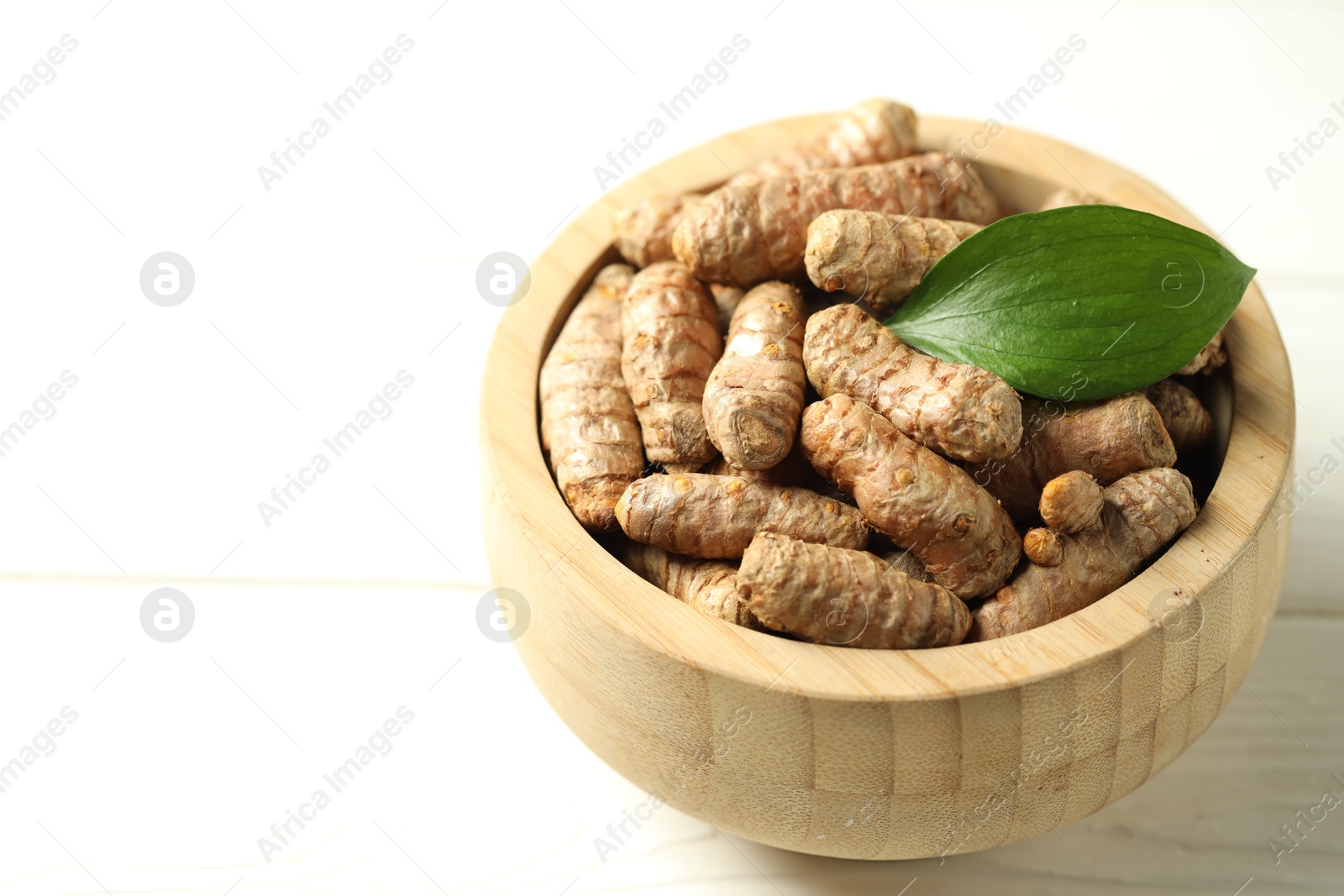 Photo of Tumeric rhizomes with leaf in bowl on white wooden table, closeup. Space for text