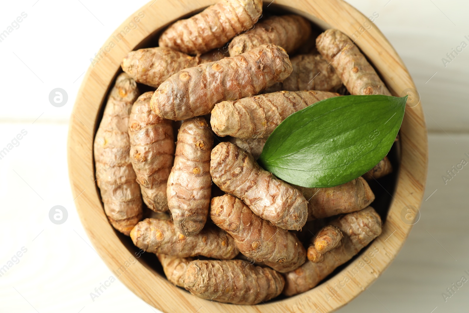 Photo of Tumeric rhizomes with leaf in bowl on white wooden table, top view