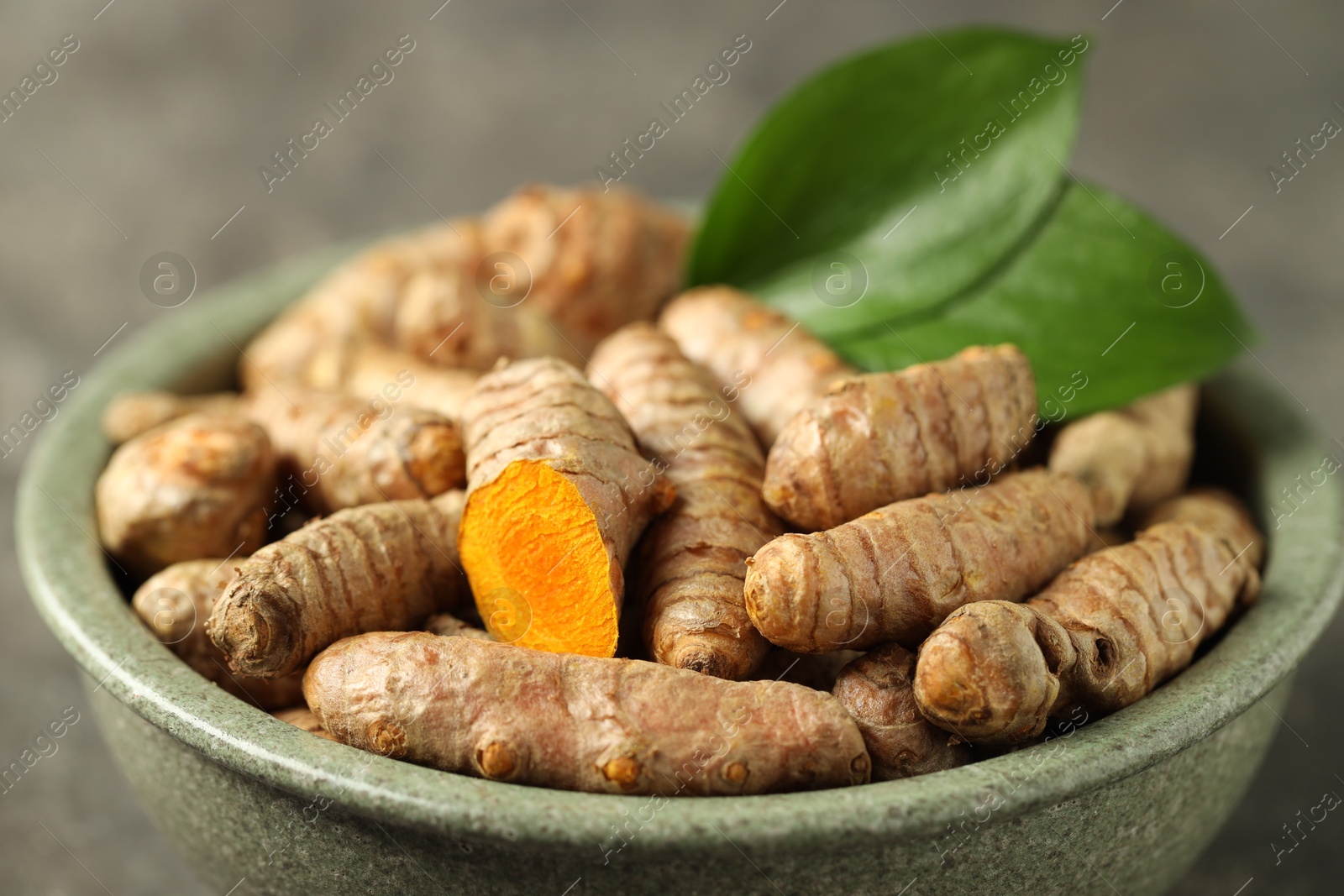 Photo of Tumeric rhizomes with leaves in bowl on grey table, closeup