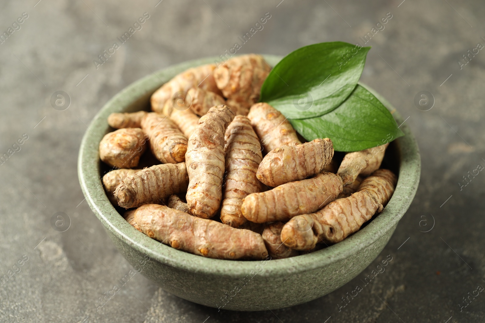 Photo of Tumeric rhizomes with leaves in bowl on grey table, closeup