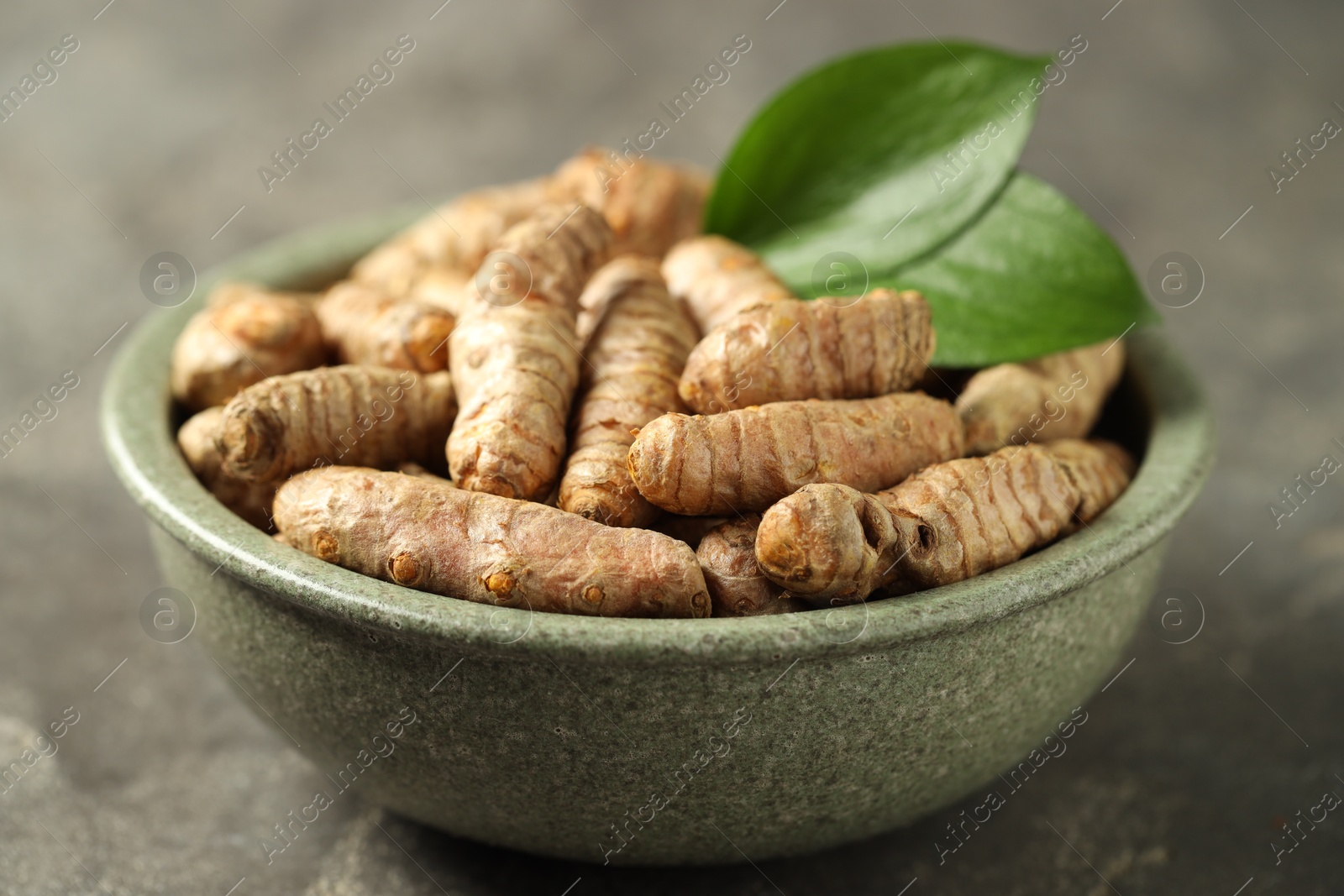 Photo of Tumeric rhizomes with leaves in bowl on grey table, closeup