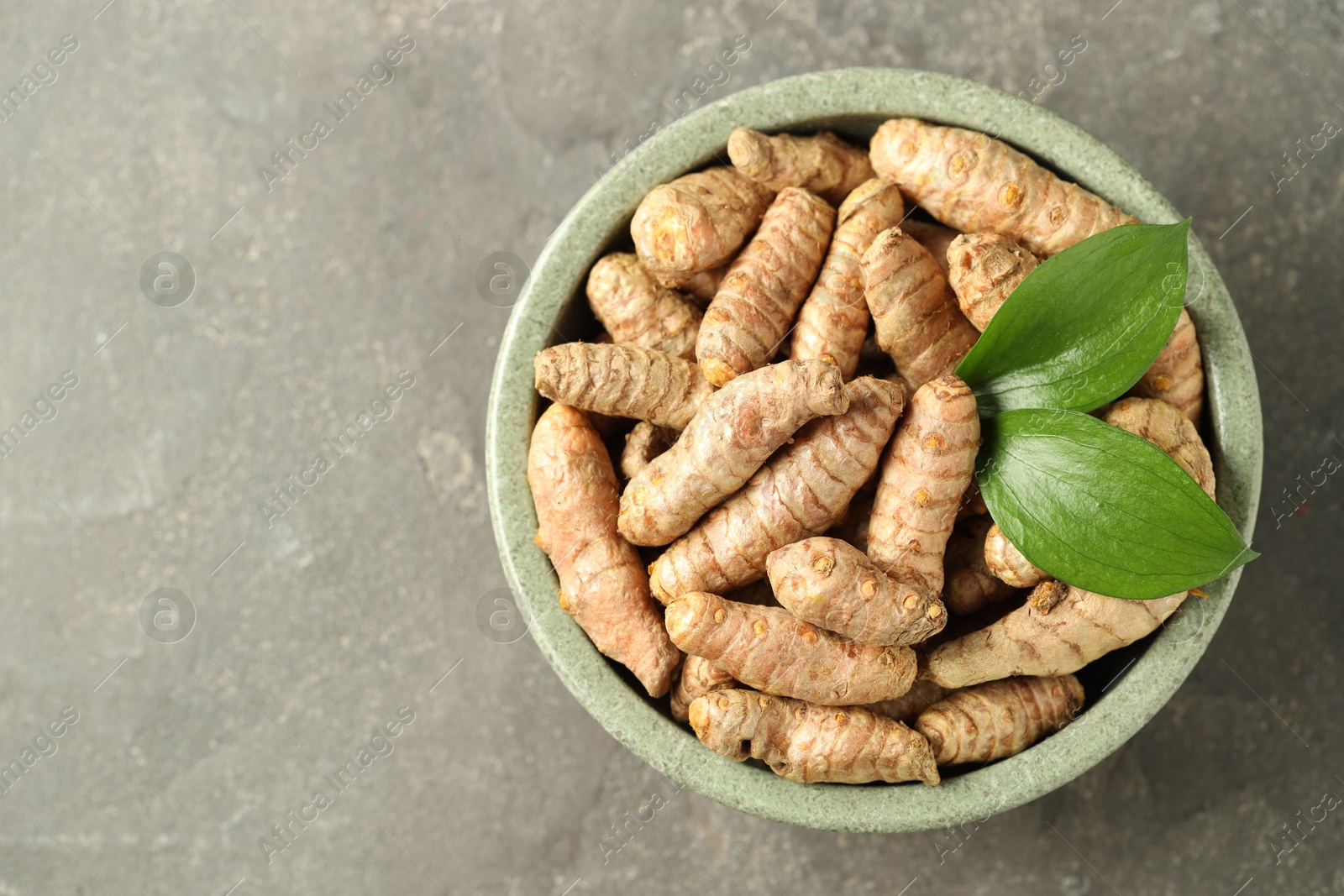 Photo of Tumeric rhizomes with leaves in bowl on grey table, top view. Space for text