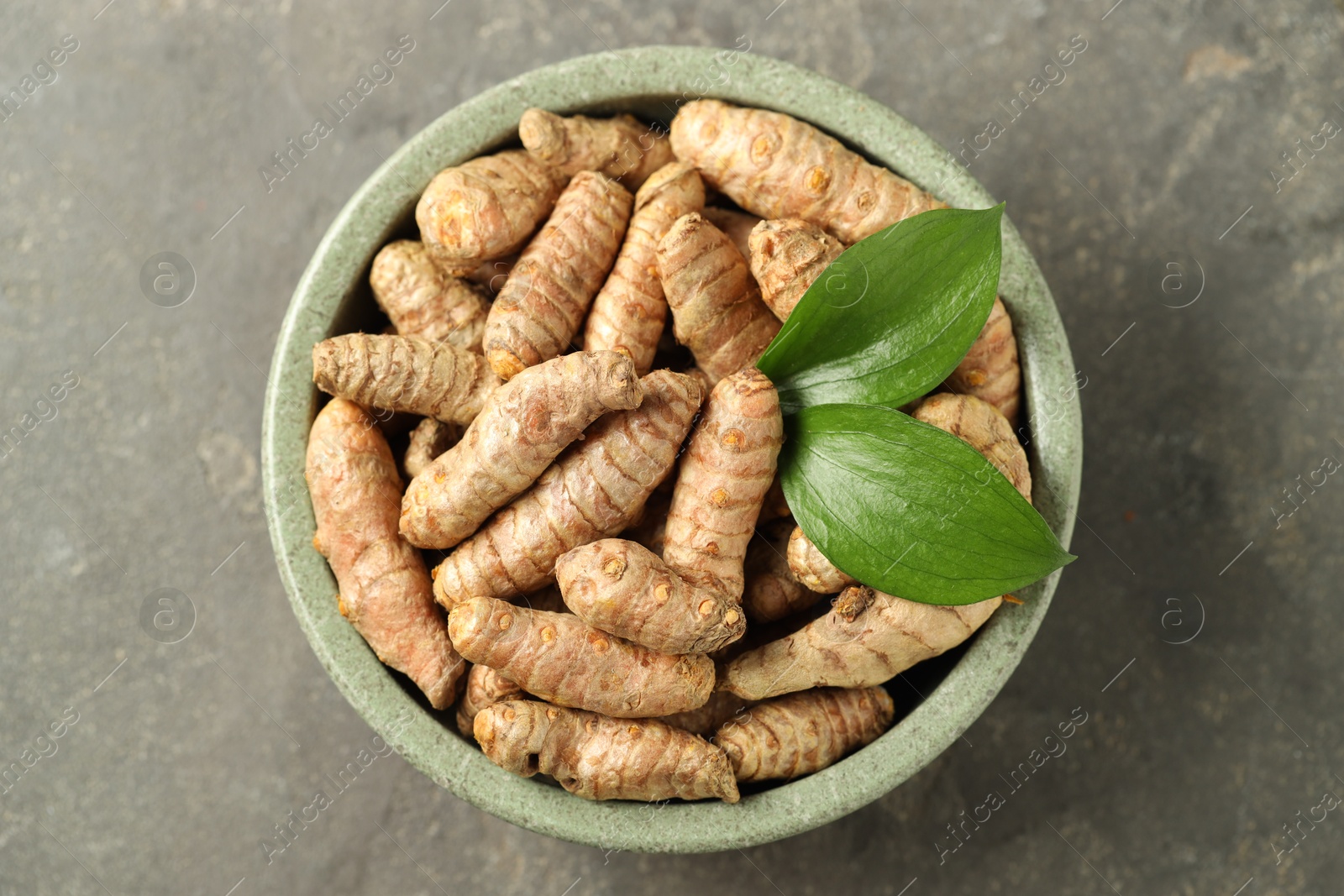 Photo of Tumeric rhizomes with leaves in bowl on grey table, top view