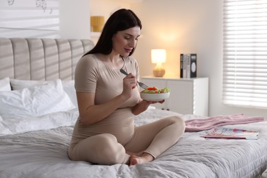 Photo of Beautiful pregnant woman eating vegetable salad on bed at home