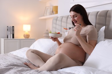 Photo of Beautiful pregnant woman eating vegetable salad on bed at home