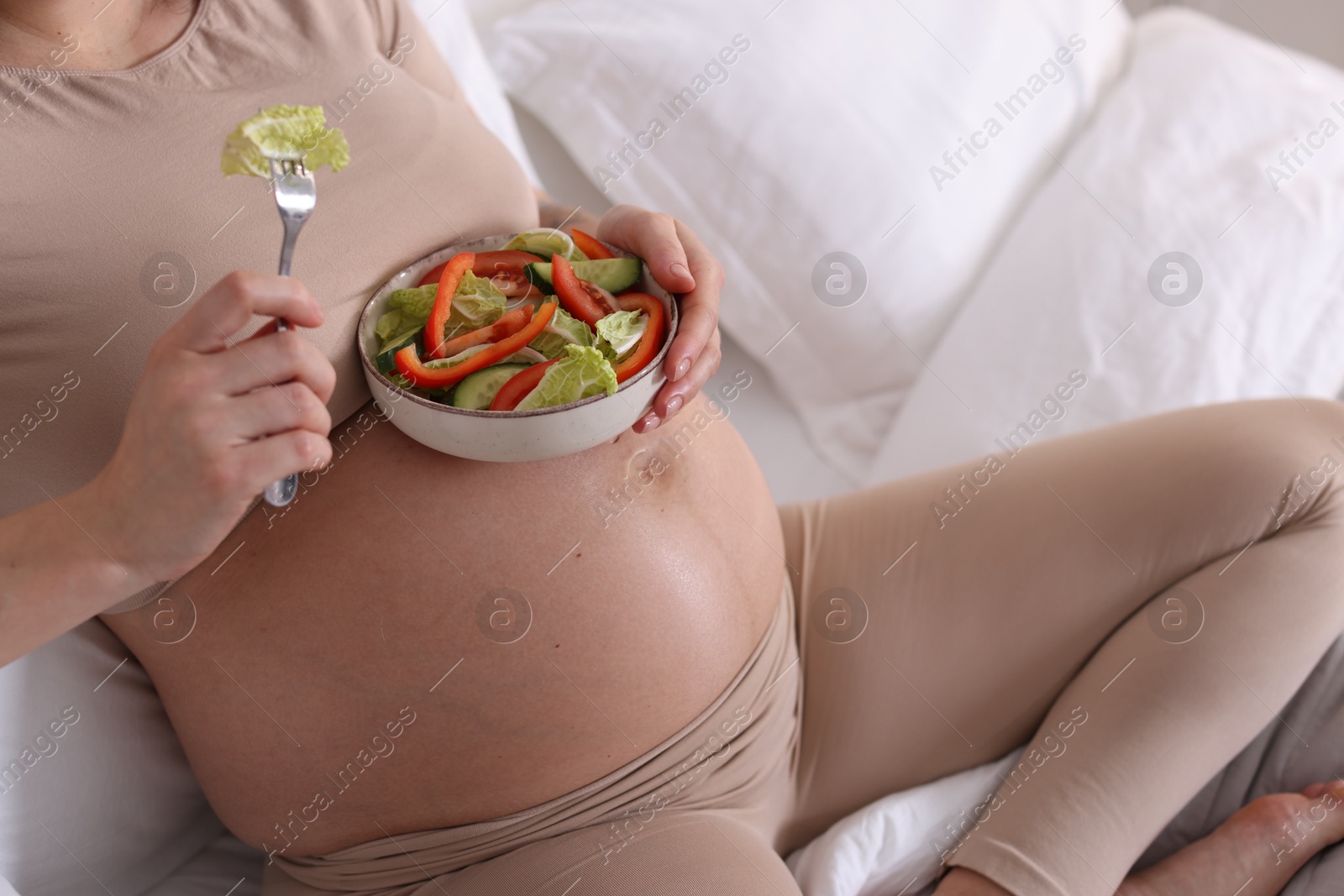 Photo of Pregnant woman eating vegetable salad on bed at home, closeup