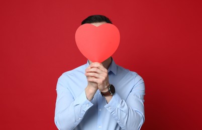 Photo of Happy Valentine's Day. Man covering his face with paper heart on red background