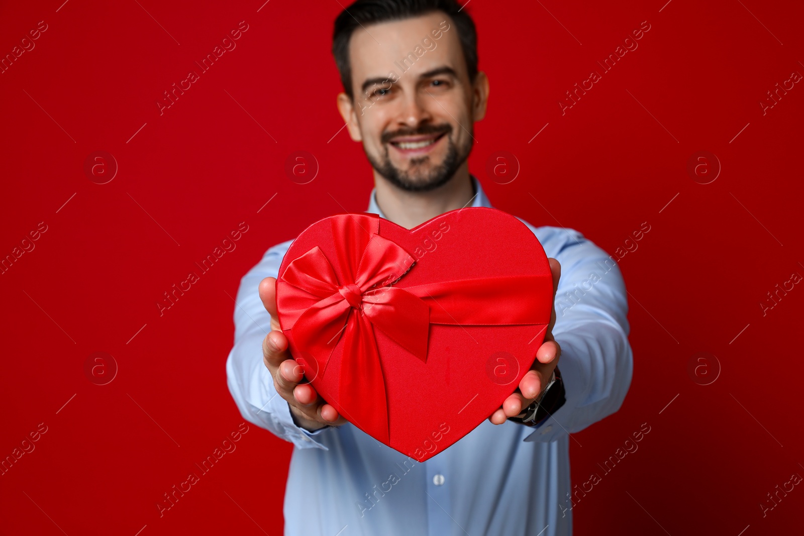 Photo of Happy Valentine's Day. Handsome man with heart shaped gift box on red background, selective focus