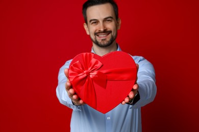 Photo of Happy Valentine's Day. Handsome man with heart shaped gift box on red background, selective focus