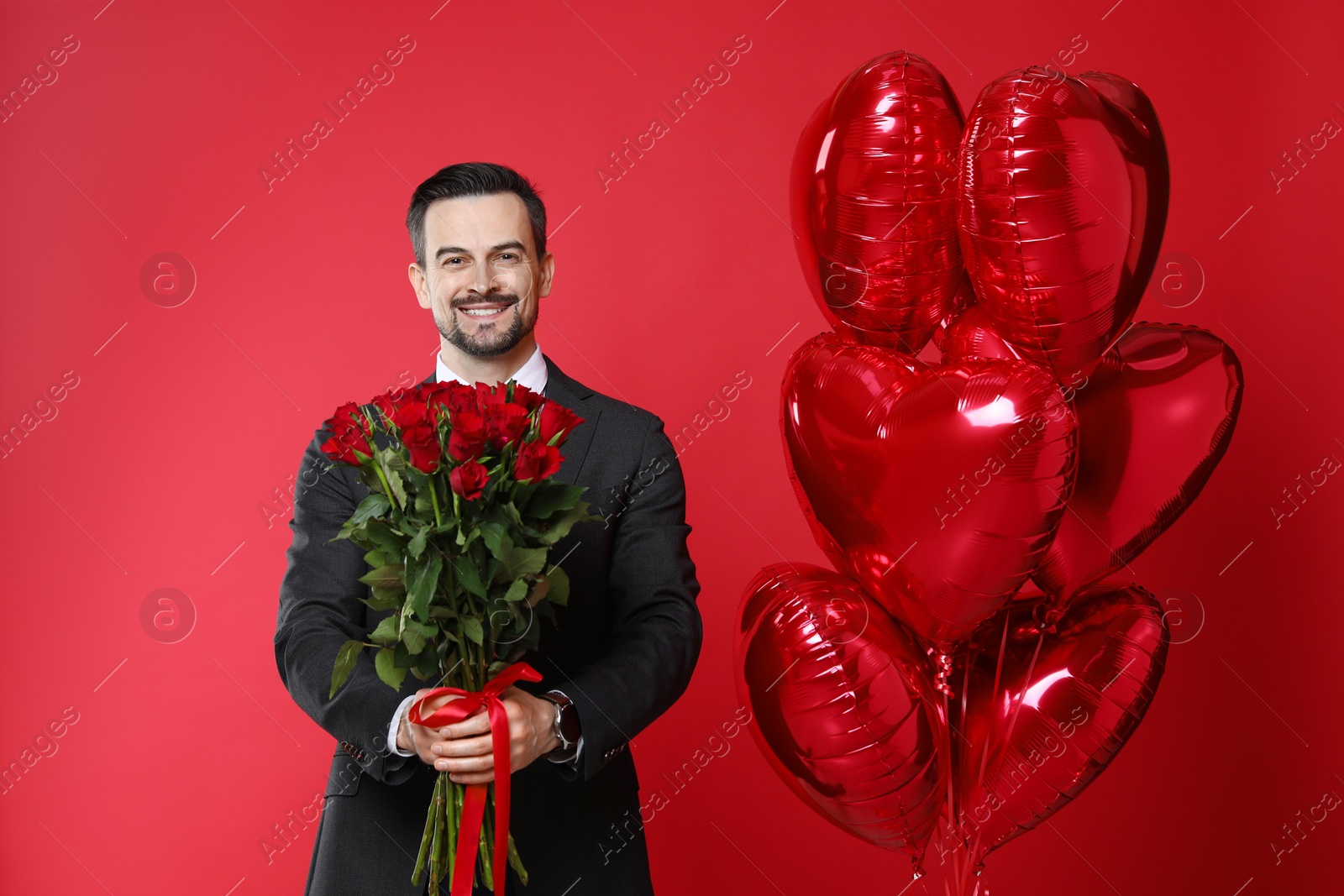 Photo of Happy Valentine's Day. Handsome man with heart shaped balloons and bouquet of roses on red background