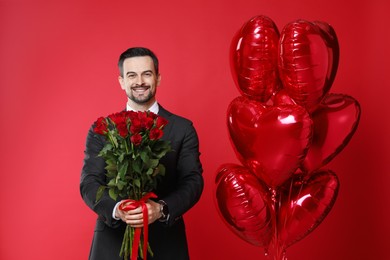 Photo of Happy Valentine's Day. Handsome man with heart shaped balloons and bouquet of roses on red background