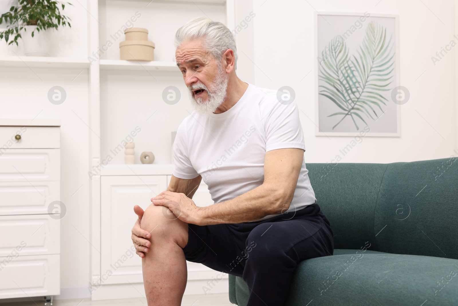 Photo of Senior man suffering from knee pain on sofa at home