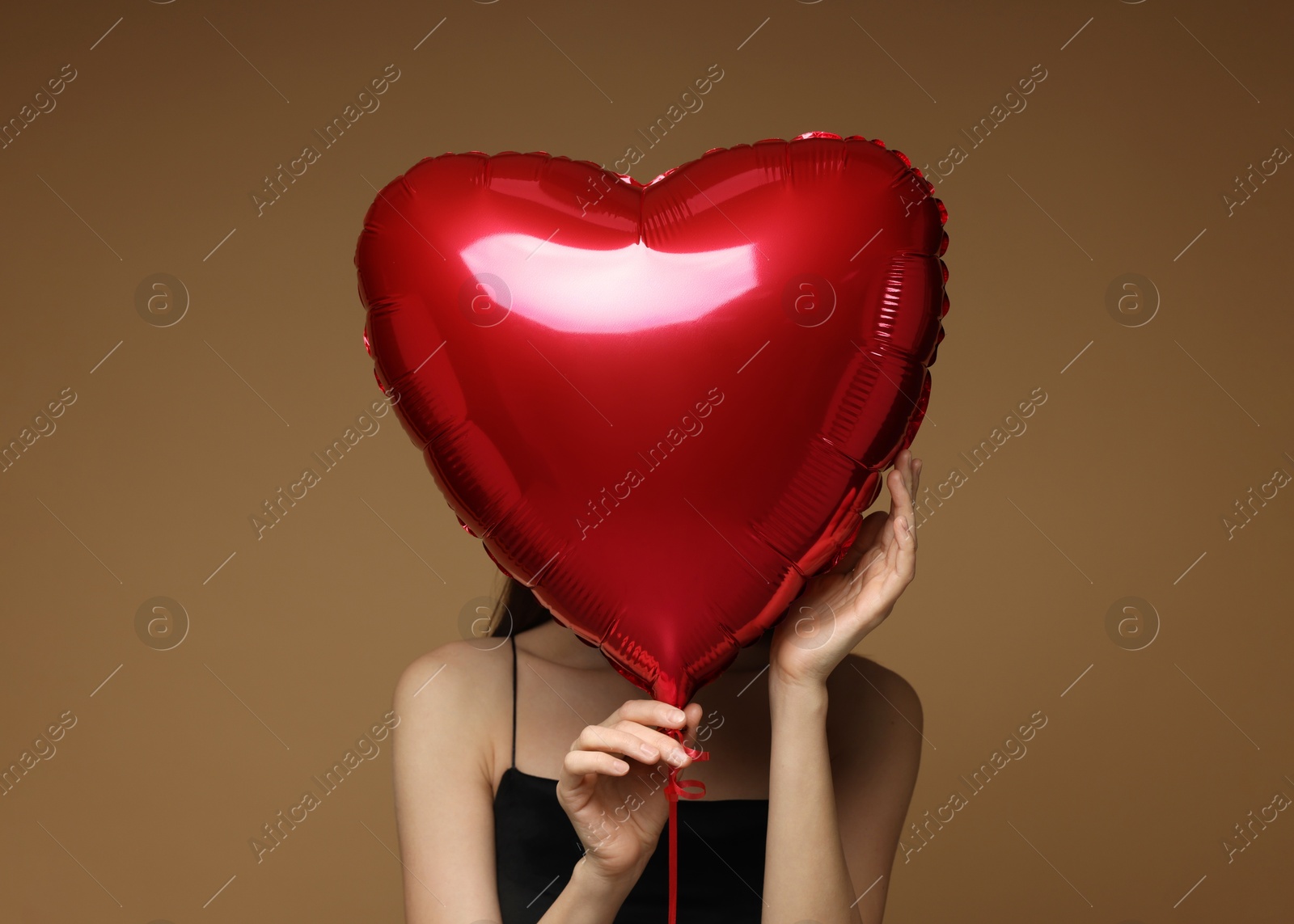 Photo of Happy Valentine's Day. Woman hiding behind heart shaped balloon on brown background