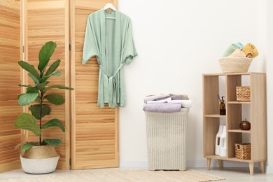 Photo of Wicker basket full of laundry, bathrobe and houseplant in bathroom