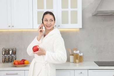 Photo of Beautiful woman with apple talking on smartphone after spa procedure in kitchen