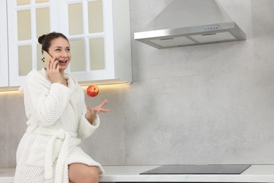 Photo of Beautiful woman throwing apple while talking on smartphone after spa procedure in kitchen