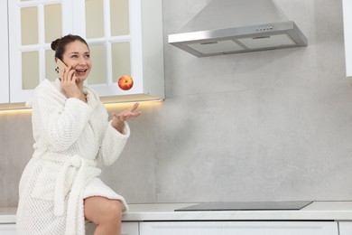 Photo of Beautiful woman throwing apple while talking on smartphone after spa procedure in kitchen