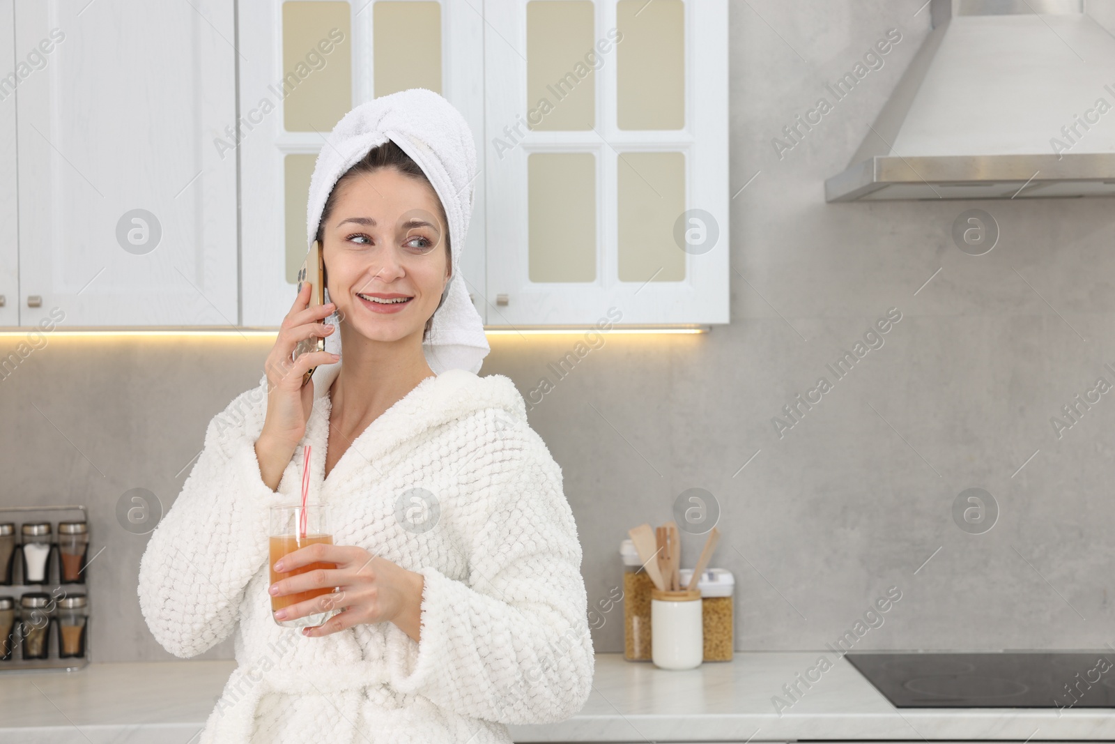 Photo of Beautiful woman with glass of juice talking on smartphone after spa procedure in kitchen, space for text