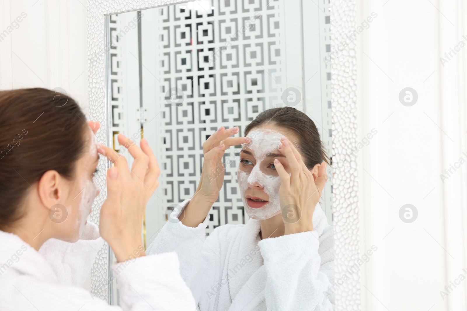 Photo of Spa day. Beautiful woman applying mask onto her face near mirror indoors