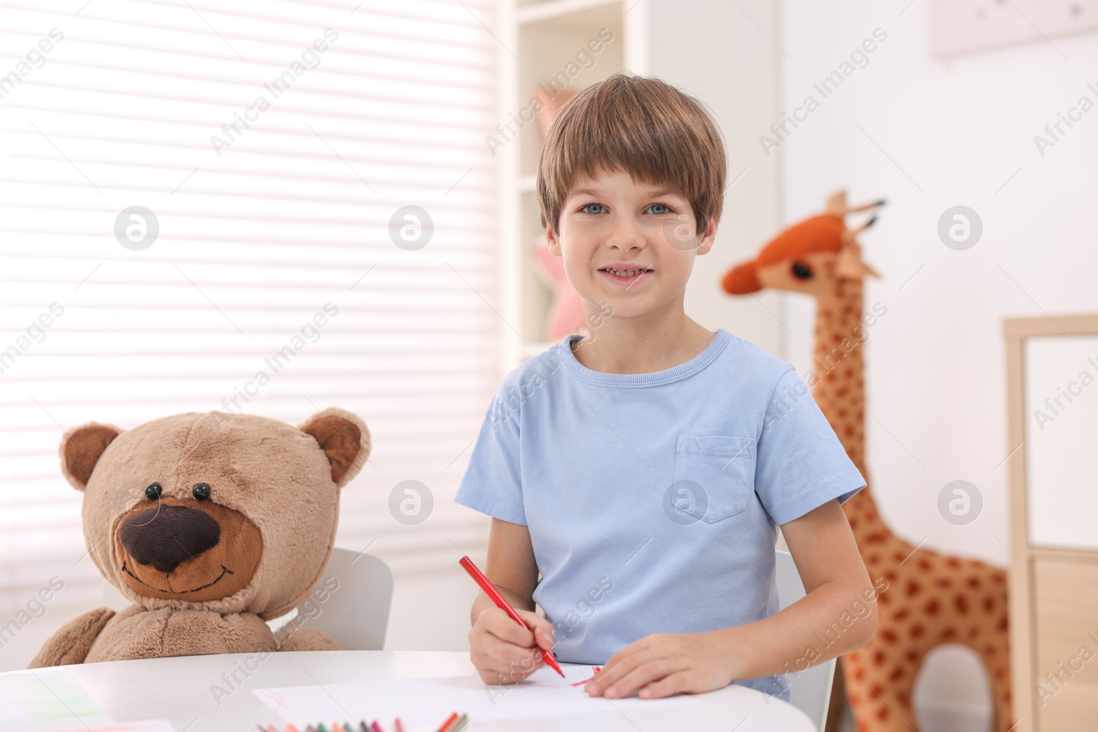 Photo of Smiling boy drawing near teddy bear at white table in kindergarten