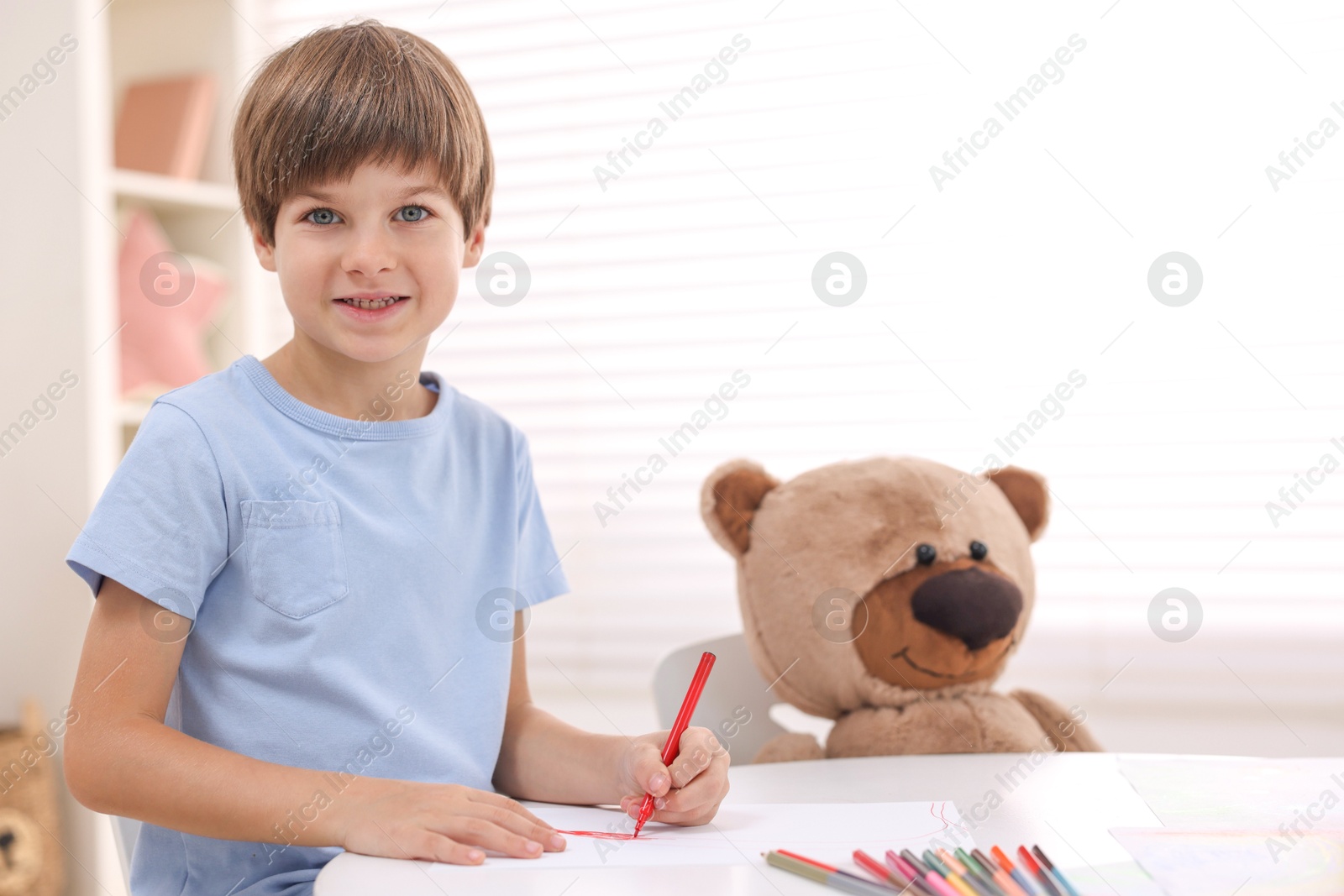 Photo of Smiling boy drawing near teddy bear at white table in kindergarten