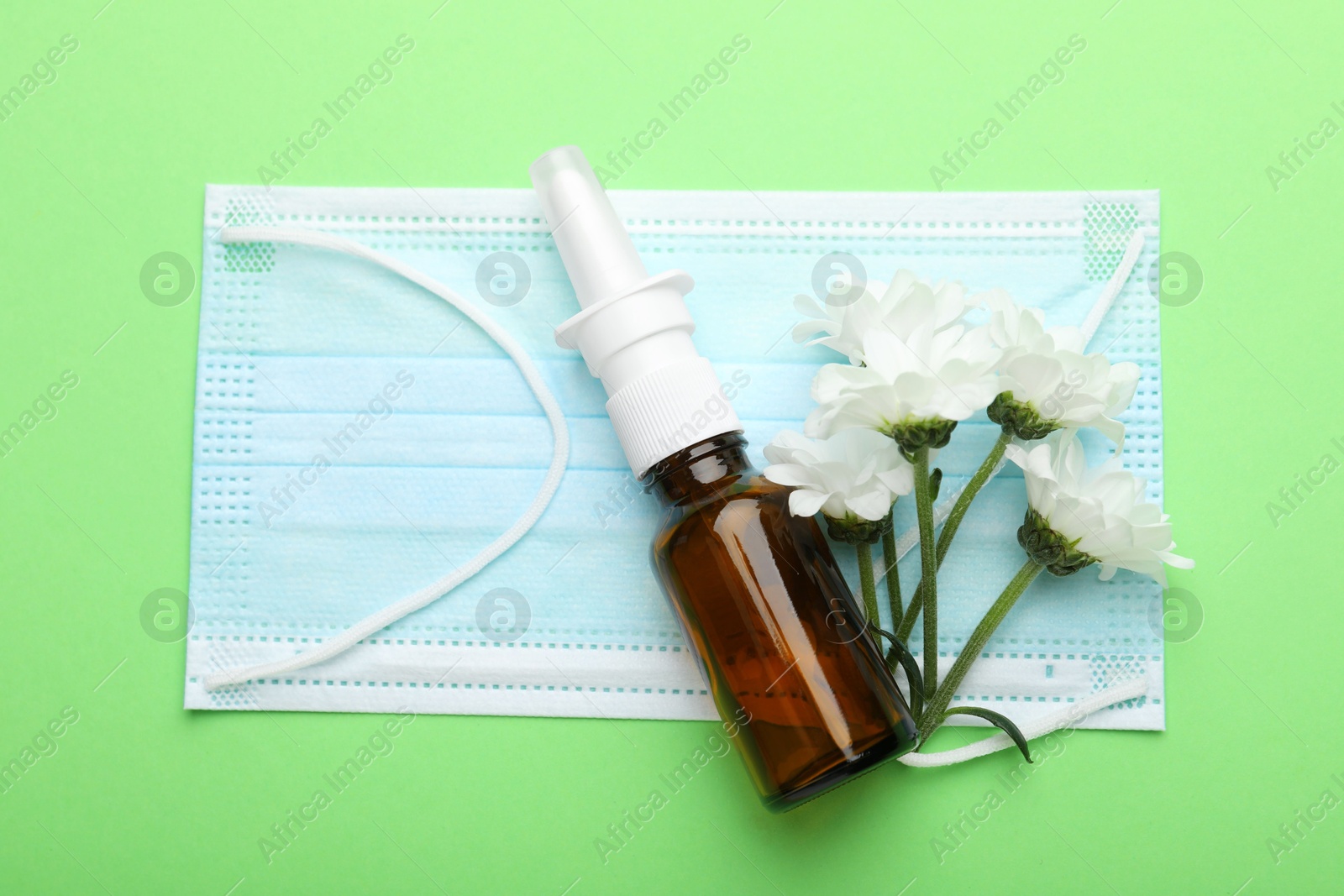 Photo of Allergy treatment. Nasal spray, protective mask and flowers on green background, top view