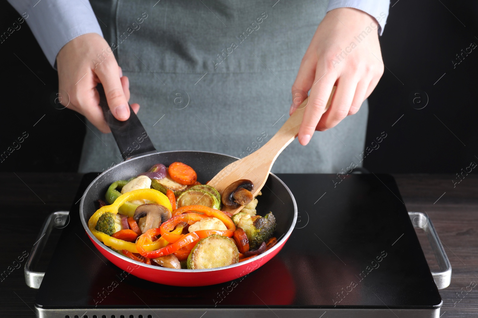 Photo of Woman frying vegetables and mushrooms in pan on stove at wooden table, closeup