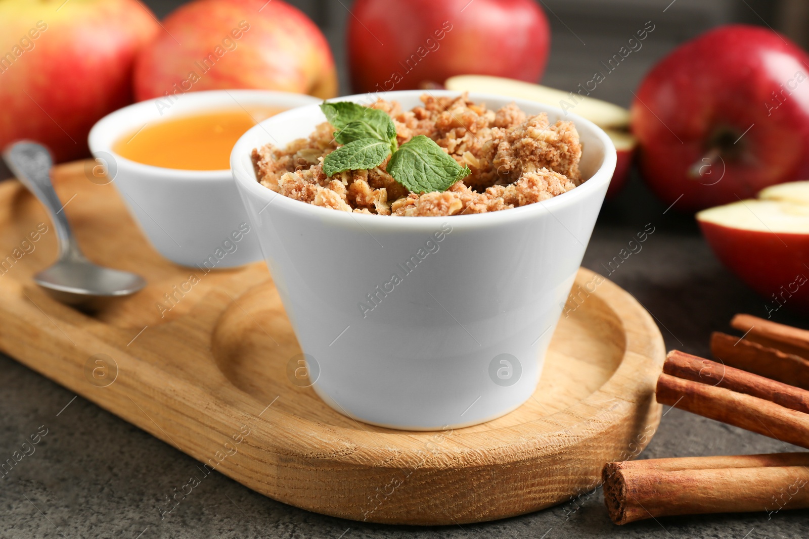 Photo of Delicious apple crisp in bowl, fresh fruits, cinnamon sticks, honey and mint on grey table, closeup