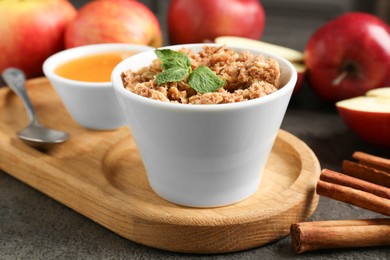 Photo of Delicious apple crisp in bowl, fresh fruits, cinnamon sticks, honey and mint on grey table, closeup