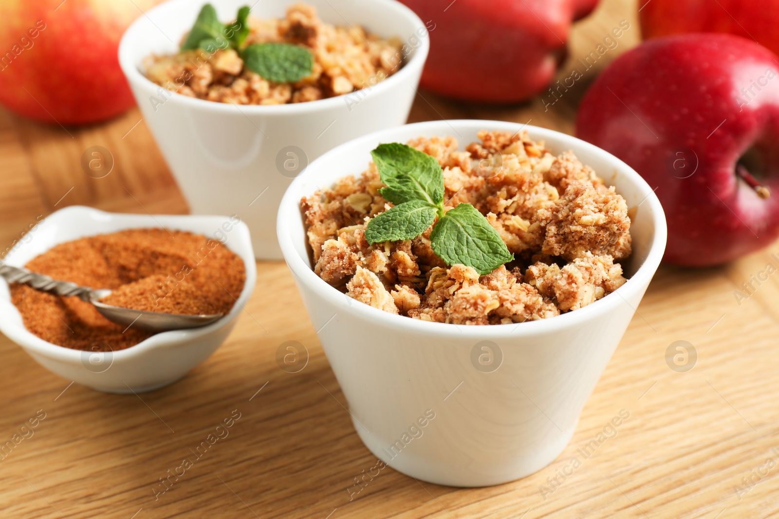 Photo of Delicious apple crisp with mint in bowls, cinnamon and fresh fruits on wooden table, closeup