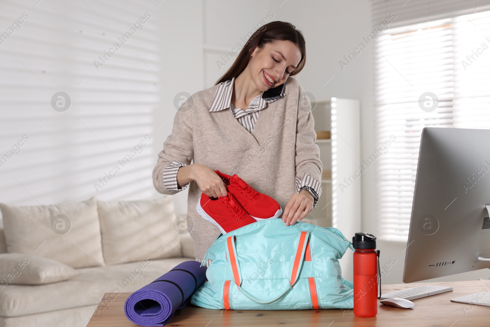 Photo of Happy woman talking on phone while packing gym bag at desk indoors