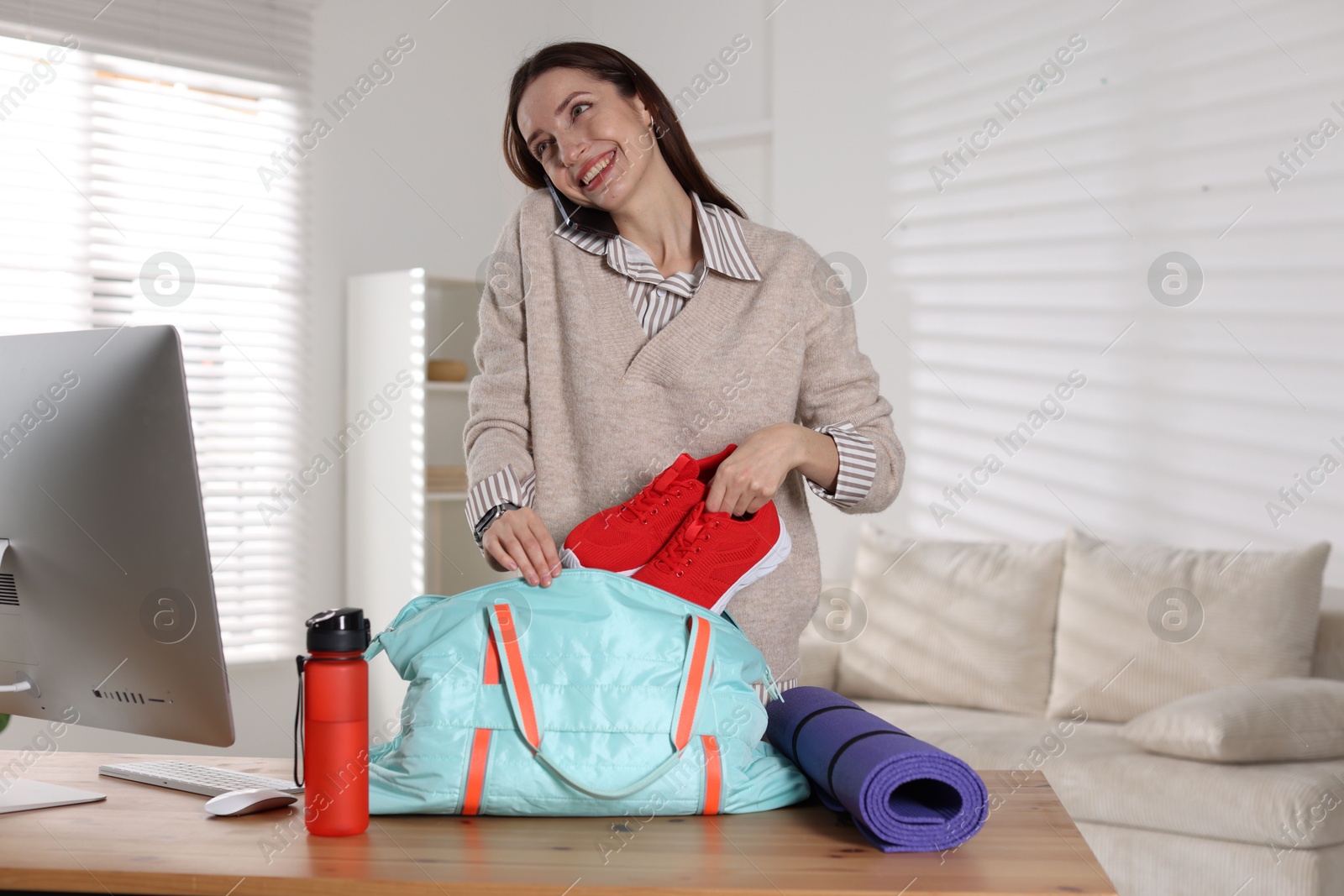 Photo of Happy woman talking on phone while packing gym bag at desk indoors