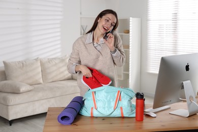 Photo of Happy woman talking on phone while packing gym bag at desk indoors